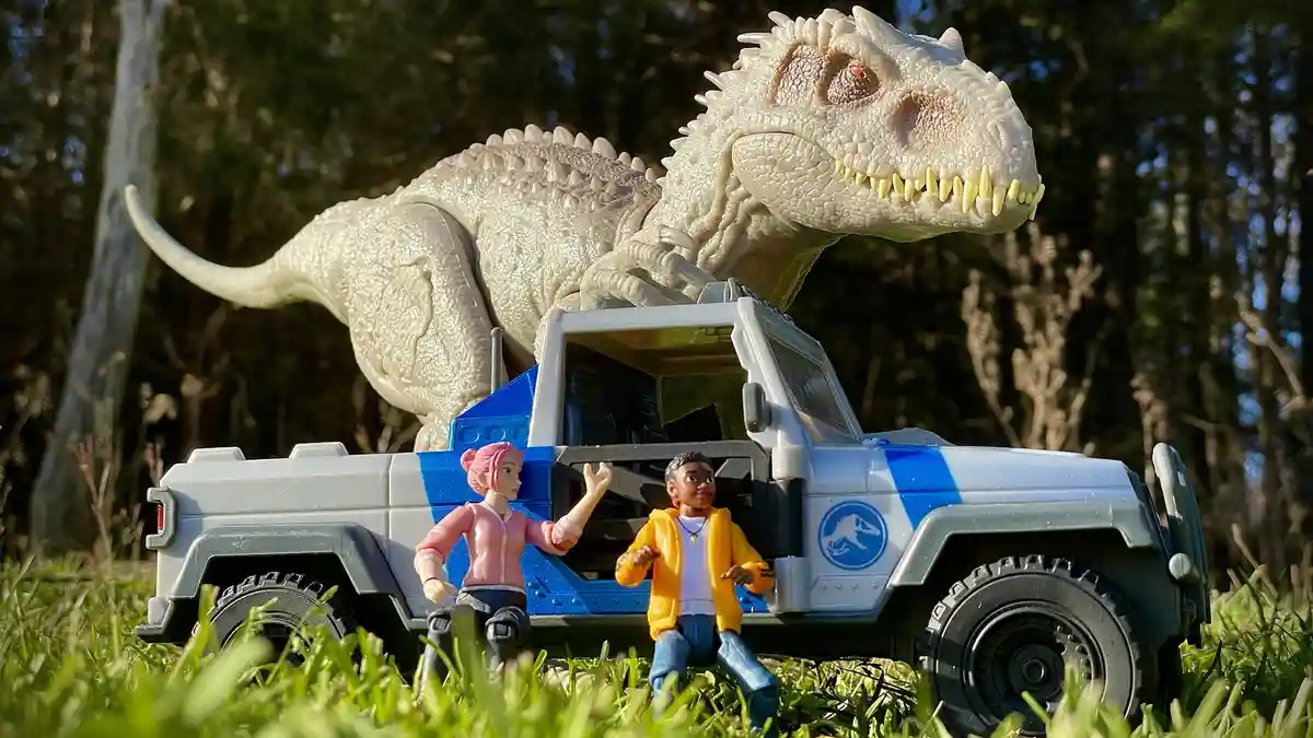 A toy scene shows two human figurines sitting beside a blue and white Jurassic Park-style jeep in a grassy outdoor setting…