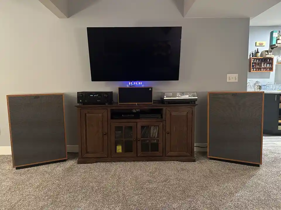 A room with a large TV mounted on a gray wall above a wooden media console. On the console are an AV receiver, a subwoofer…
