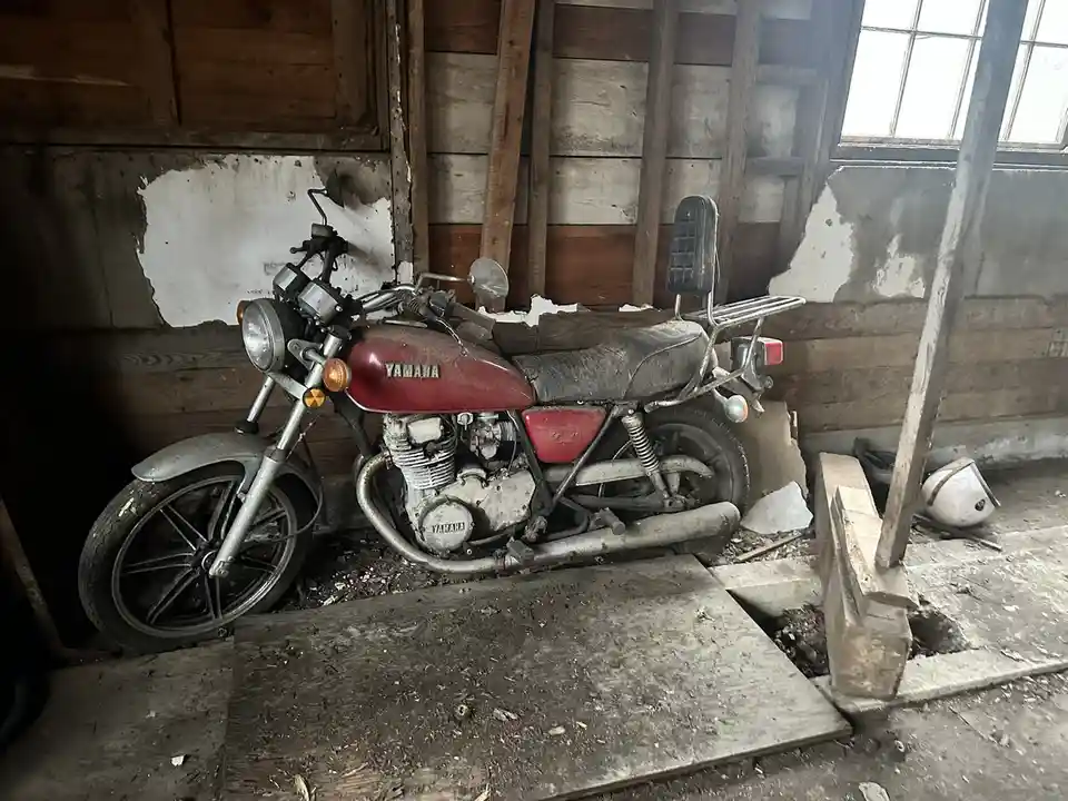 A red Yamaha motorcycle sits abandoned and dusty inside a dilapidated wooden shed with peeling paint and debris. The bike is…