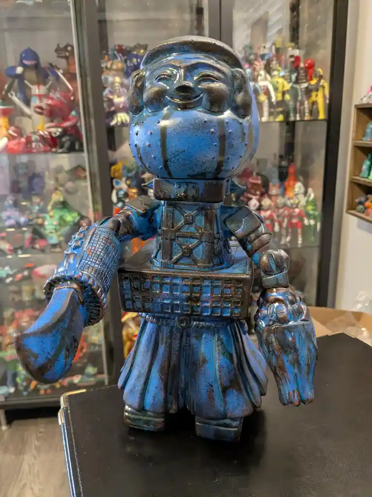 A blue, rusted, robot-like figurine with a smiling, human-like face and ornate armor stands on a black surface. Behind it, a…