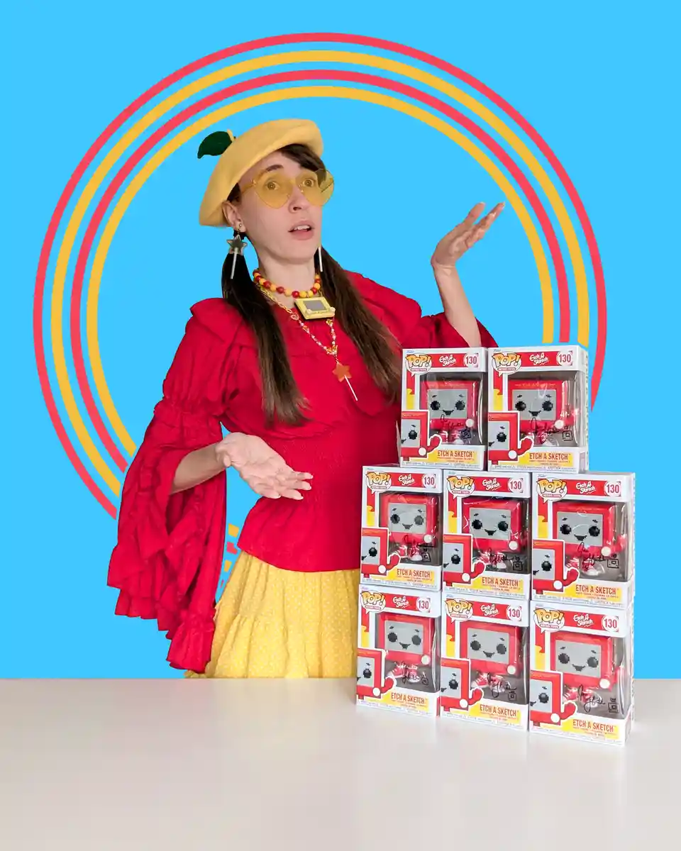 A woman in a red ruffled top, yellow polka-dot skirt, and a yellow beret with a green leaf stands behind a table piled with…