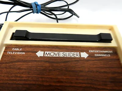 A close-up of a vintage wooden cable box with a black slider handle. The box has engraved text: 'CABLE TELEVISION' on the…