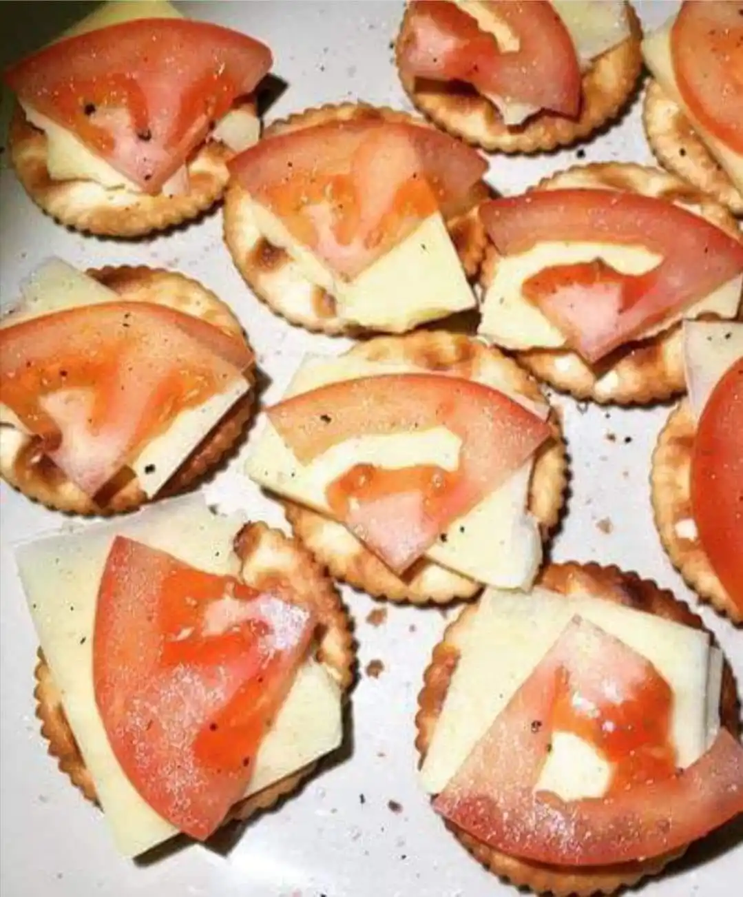 A close-up photo shows multiple round crackers topped with slices of tomato and cheese, seasoned with black pepper, arranged…