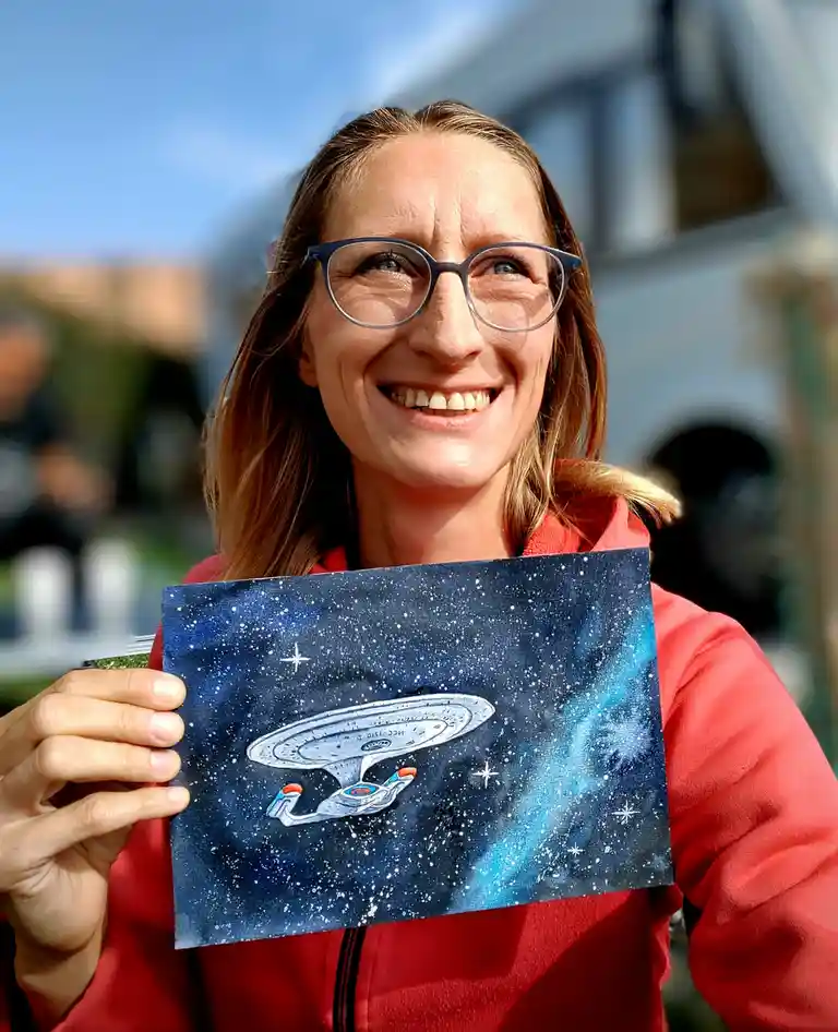 A smiling woman with glasses and a red jacket holds up a painting of the Starship Enterprise from Star Trek against a…
