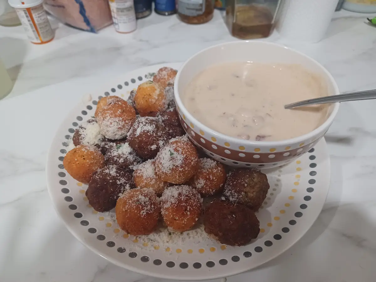 A plate of golden-brown fried balls, dusted with grated cheese and herbs, sits next to a bowl of creamy dipping sauce. The…