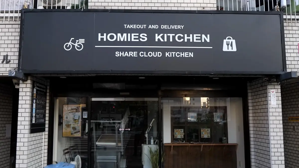 A storefront with a black sign that reads 'TAKEOUT AND DELIVERY HOMIES KITCHEN SHARE CLOUD KITCHEN', flanked by a bicycle…