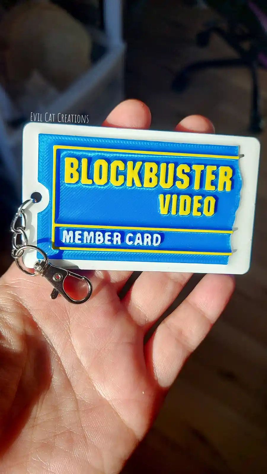 A hand holds a small, 3D-printed keychain replica of a classic Blockbuster Video member card, featuring the iconic blue and…