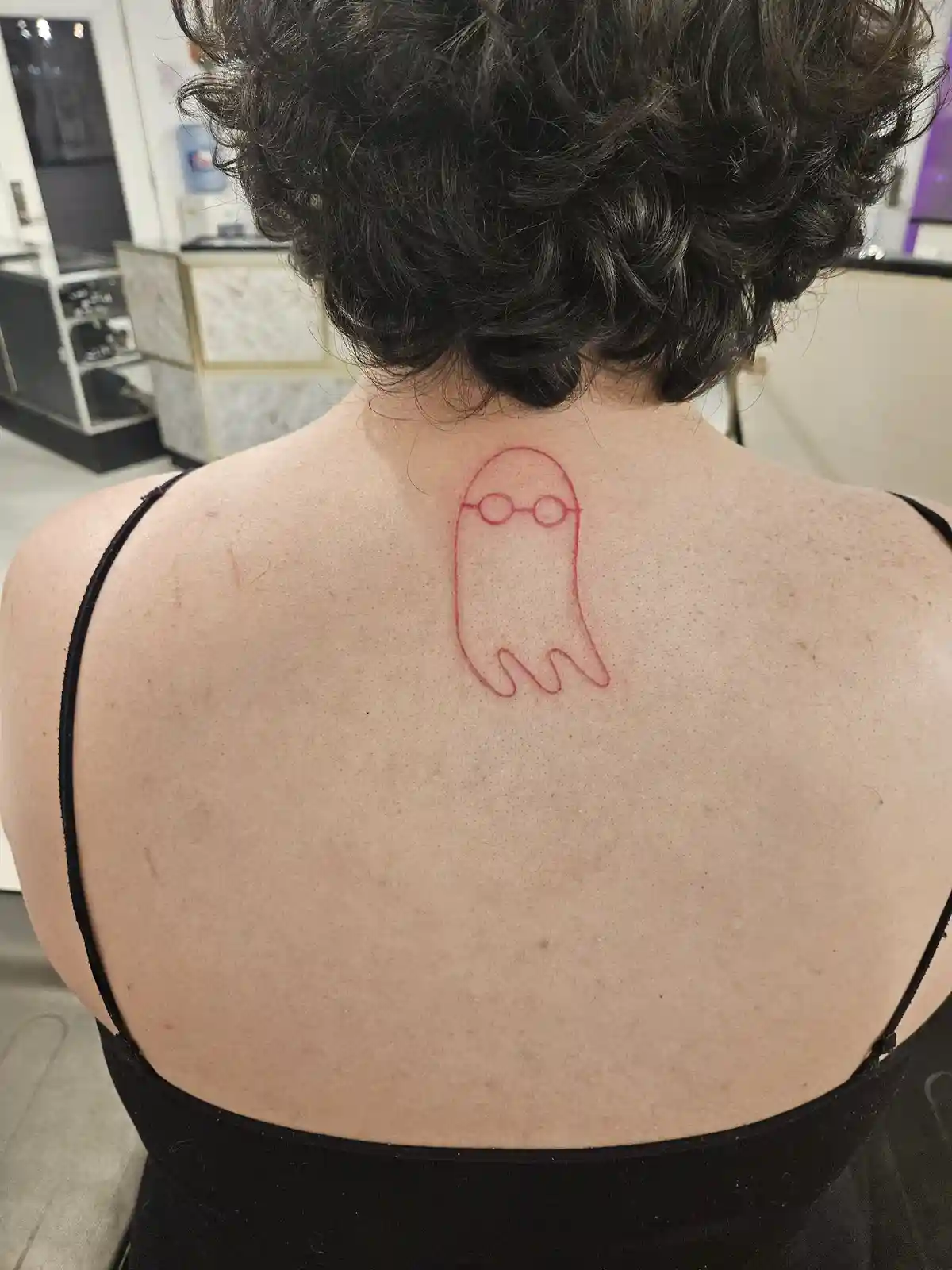 The image shows the back of a person with short dark curly hair, wearing a black top, revealing a red outline tattoo of a…