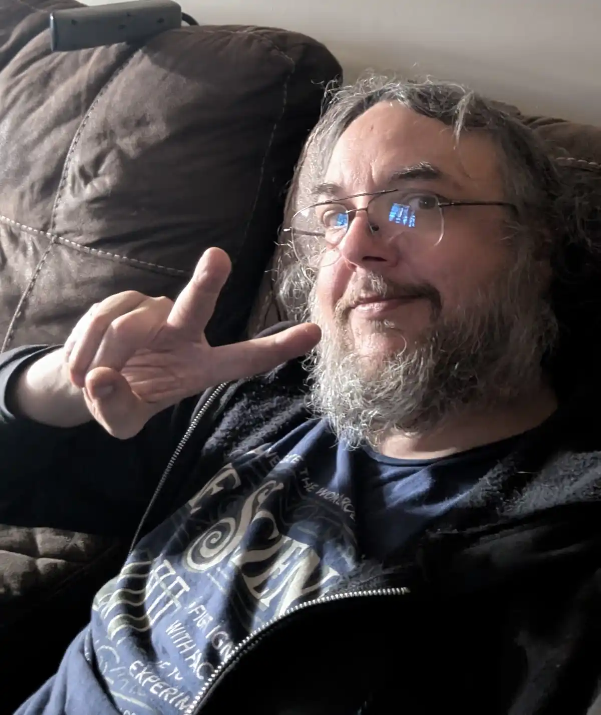 A man with a gray beard and glasses, wearing a black zip-up hoodie over a blue t-shirt, sits on a brown couch and makes a…
