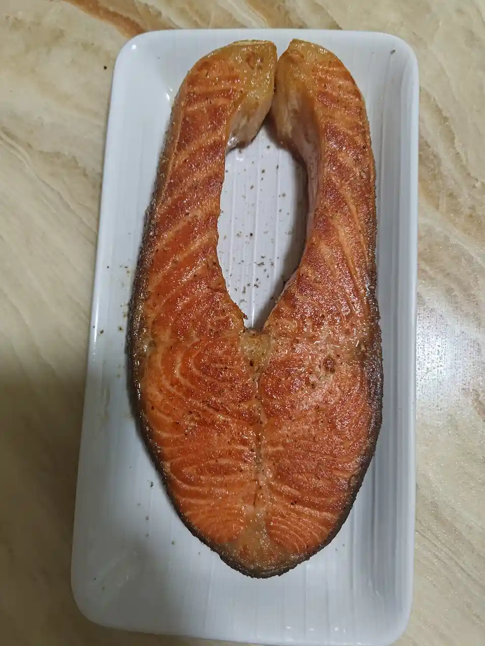 A cooked salmon fillet sits on a white rectangular plate, its shape resembling the letter 'U' due to the natural curve of…