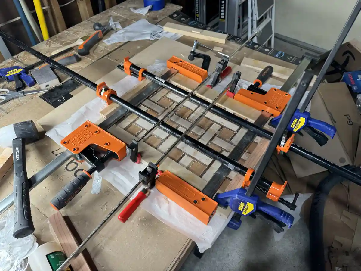 A woodworking project is heavily secured with multiple orange and blue clamps, including long bar clamps and quick-grip…