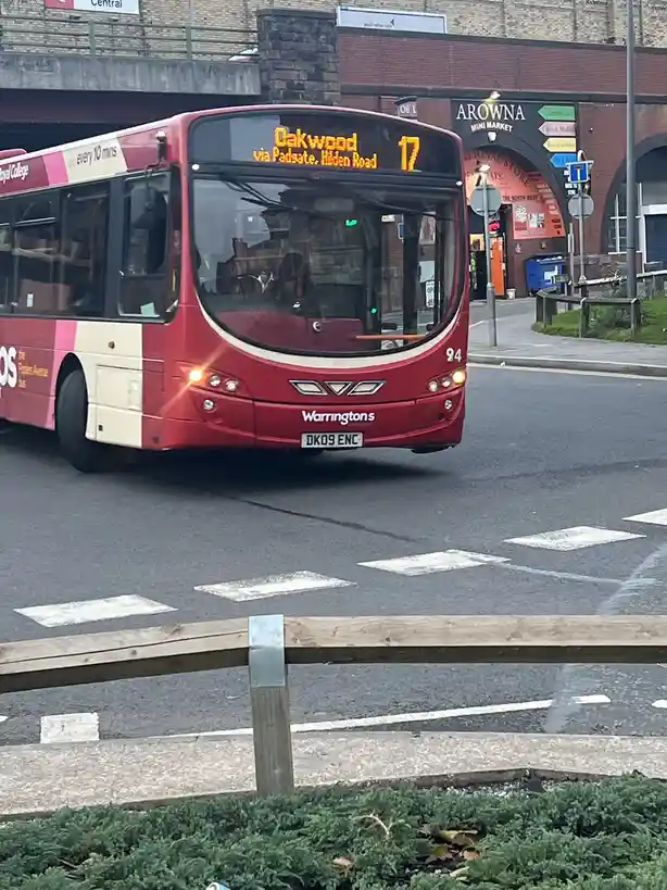A red and cream bus, route 17 to Oakwood via Padgate and Hilden Road, is captured driving on a city street. The bus…
