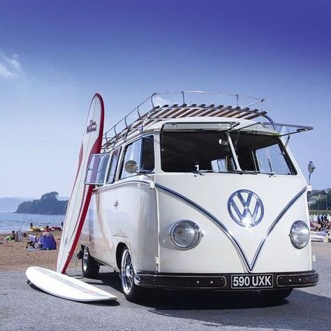 A vintage white Volkswagen Type 2 van with a surfboard mounted vertically on its side and another lying on the ground beside…