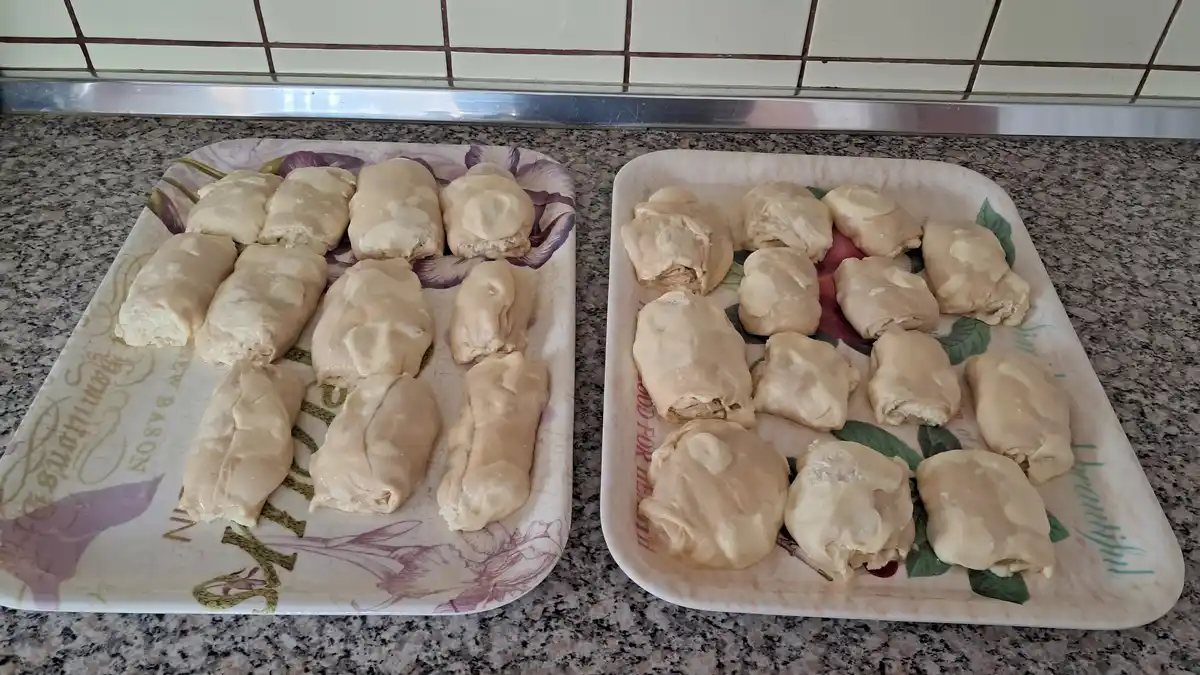 Two trays of uncooked, messy dough rolls sit side-by-side on a granite countertop, one appearing slightly more neatly formed…
