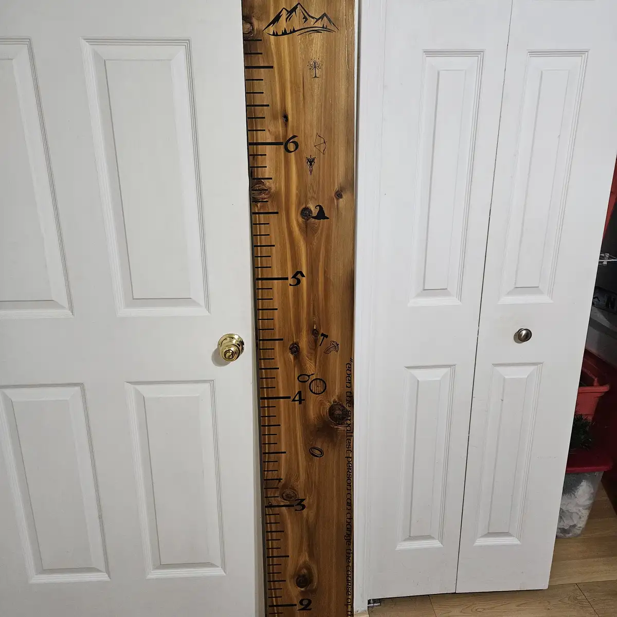 A wooden growth chart ruler stands vertically between two white doors, marked with numbers 2 through 6 and decorated with…