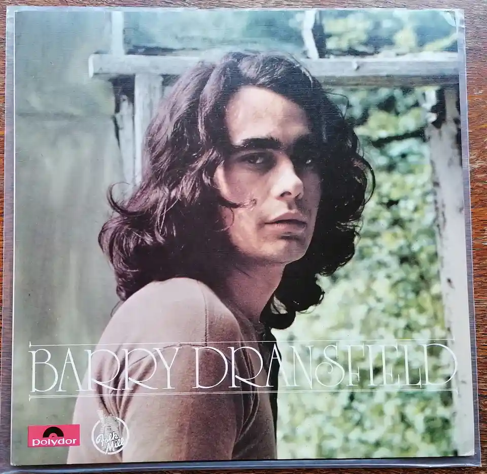 The image shows a vintage album cover featuring a man with long, wavy dark hair looking off to the side, wearing a…