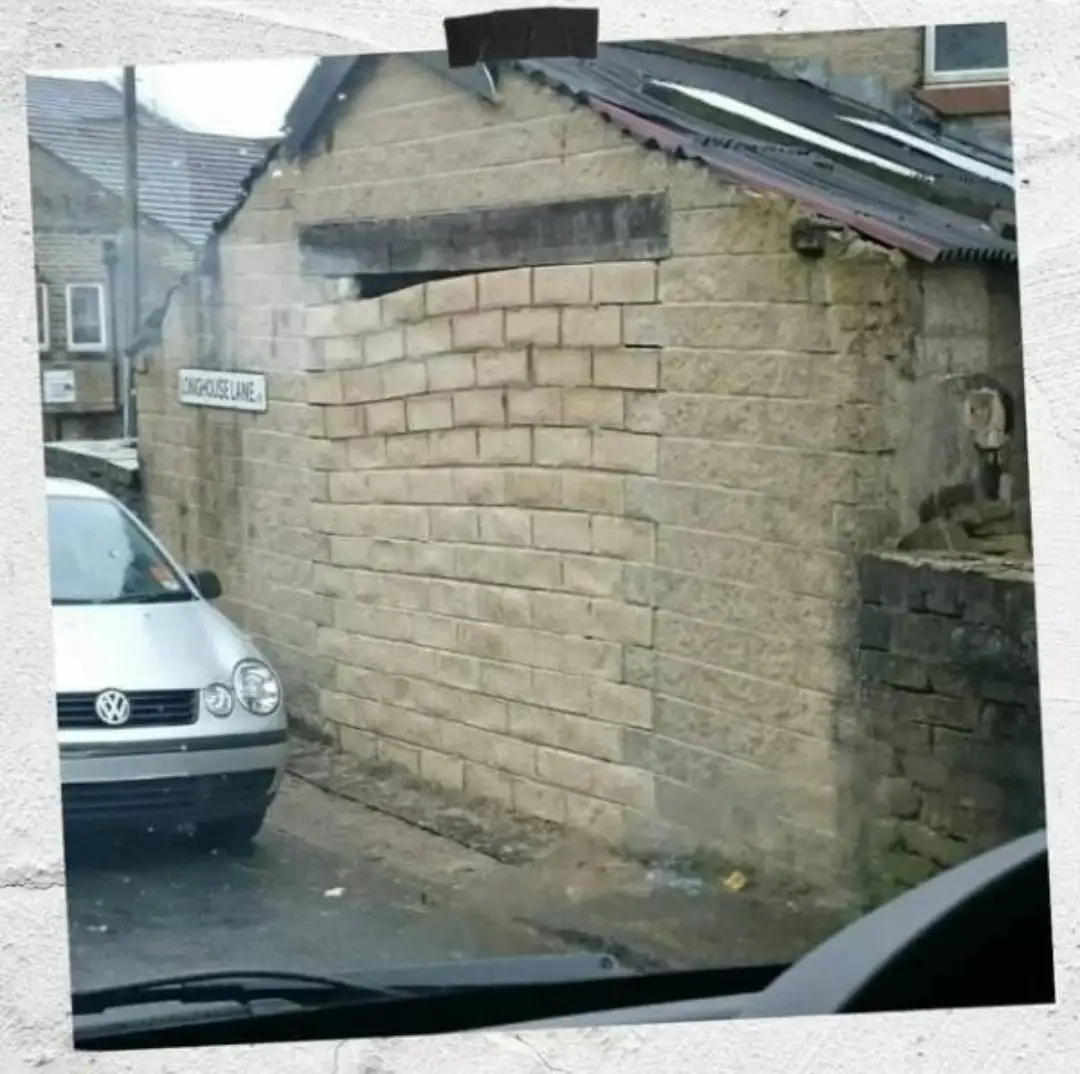 A photo taken from inside a car shows a narrow street with a brick wall on the right and a silver Volkswagen parked on the…