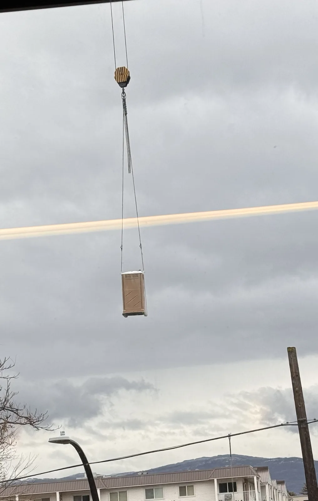 A portable toilet is suspended in mid-air by a crane's hook against a cloudy sky, with residential buildings and power lines…