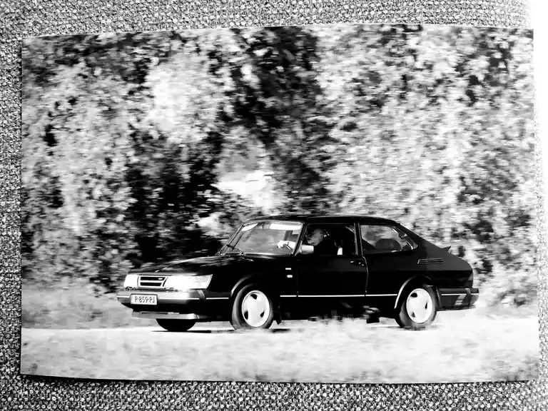 A black-and-white photograph shows a black Saab 900 driving on a road with blurred trees in the background, indicating…