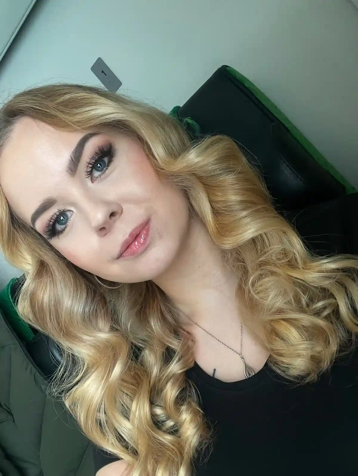 A woman with long blonde wavy hair, blue eyes, and heavy makeup including thick eyelashes and defined eyebrows, is looking…