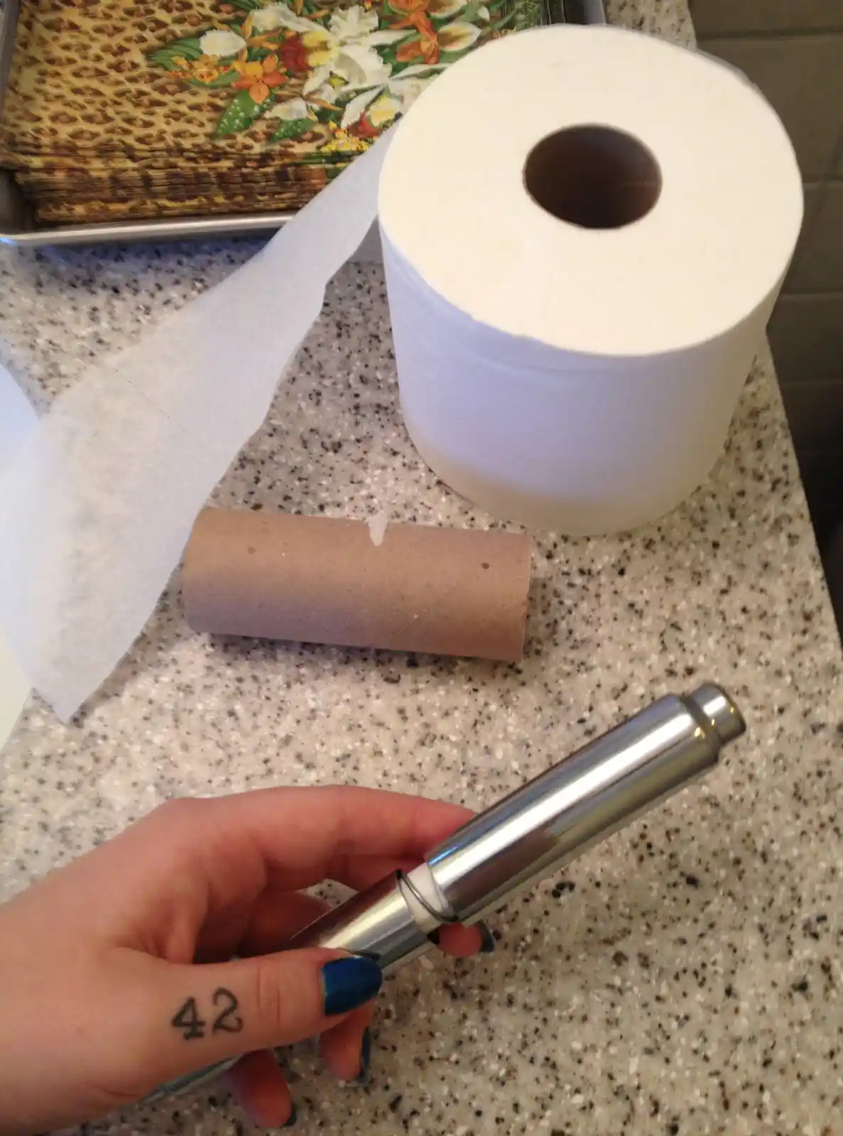A hand with blue nail polish holds a chrome toilet paper dispenser, with a full roll and empty cardboard tube on a speckled…