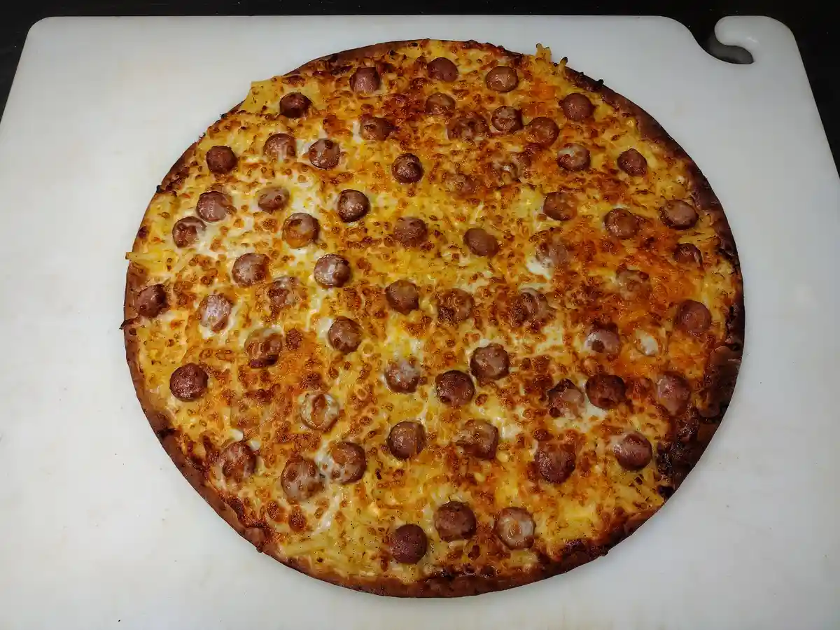 A round pizza with melted cheese and evenly distributed sausage pieces sits on a white cutting board, visually resembling a…