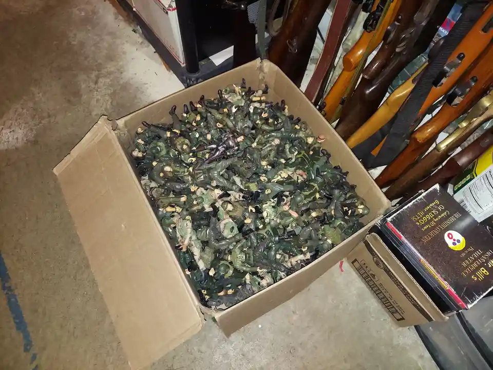 A large cardboard box overflowing with green and tan plastic army men toys sits on a concrete floor, next to a rack of real…