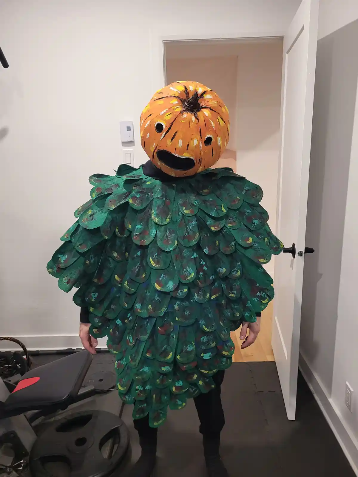 A person stands indoors wearing a homemade costume with a painted pumpkin head featuring black eyes and a wide open mouth…