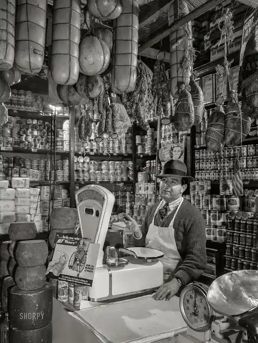 A black-and-white photo of a man in a hat and apron standing behind a counter in a vintage deli or meat shop, surrounded by…