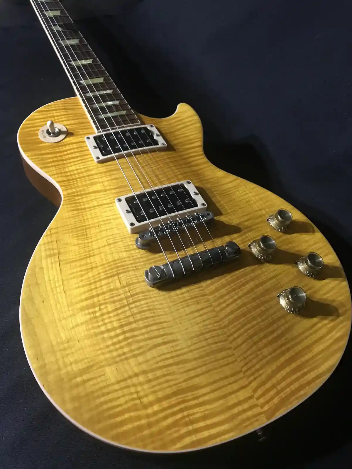 A close-up photo of a yellow electric guitar with a distinctive wood grain finish, featuring two humbucker pickups, four…