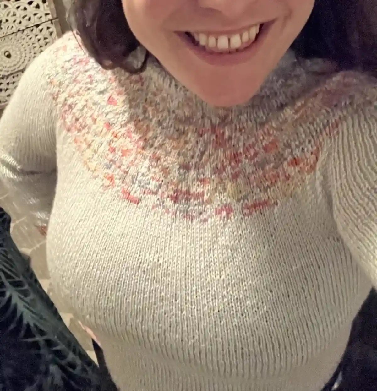 A close-up selfie shows a person smiling broadly, wearing a light-colored knit sweater with a colorful, intricate pattern…