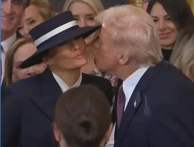 a man with blond hair and a suit kisses a woman wearing a black hat with a white band on the cheek; the woman has a neutral…