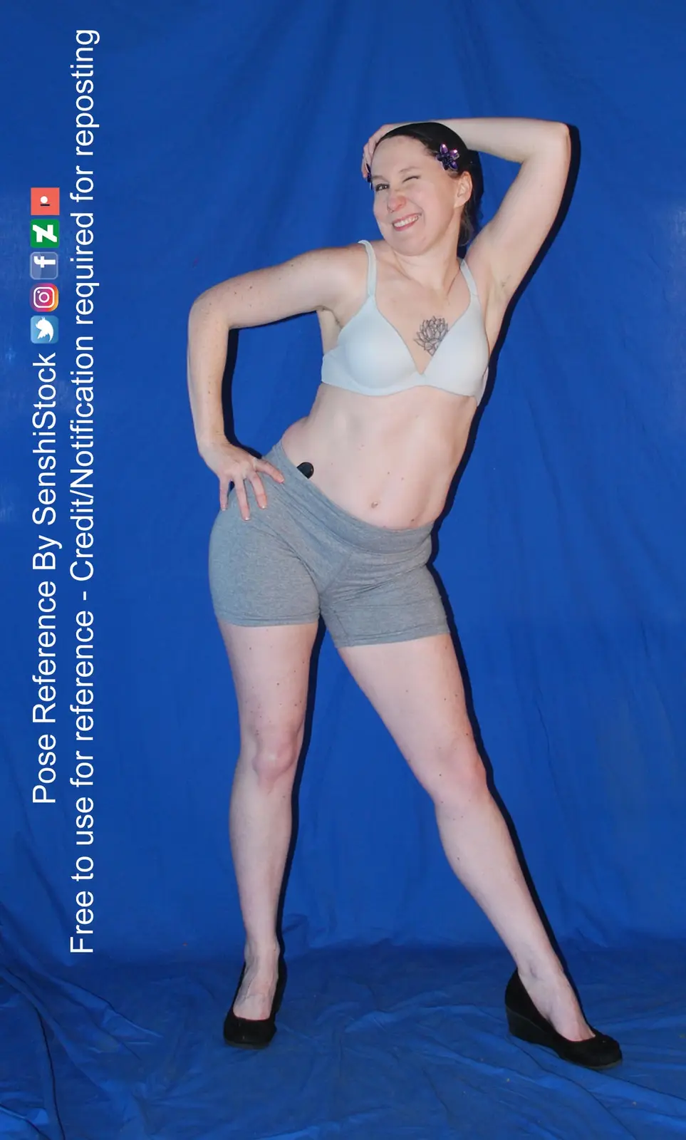 A woman in a light gray bra and gray shorts poses confidently against a solid blue backdrop, one hand on her hip and the…