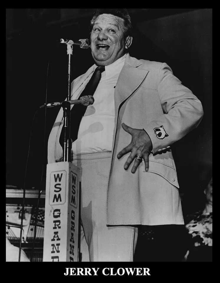 A black-and-white photo of Jerry Clower, a comedian, standing on stage in a light-colored suit with a tie, smiling broadly…
