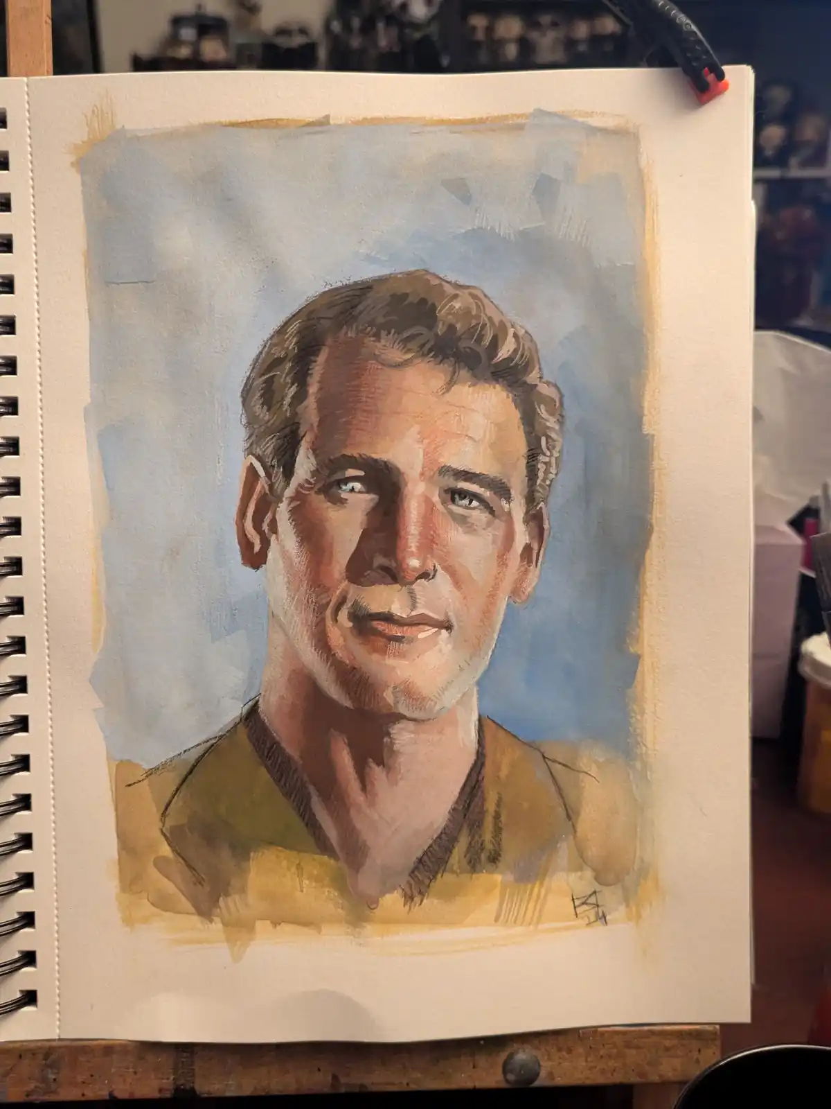 A watercolor portrait of a man with brown hair and a yellow shirt is drawn in a spiral-bound sketchbook. The text overlaid…