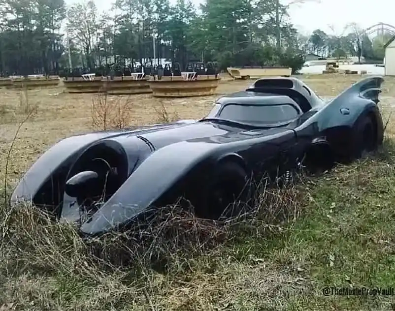 A black, bat-shaped movie prop car, resembling the Batmobile from the 1989 Batman movie, is shown abandoned and partially…