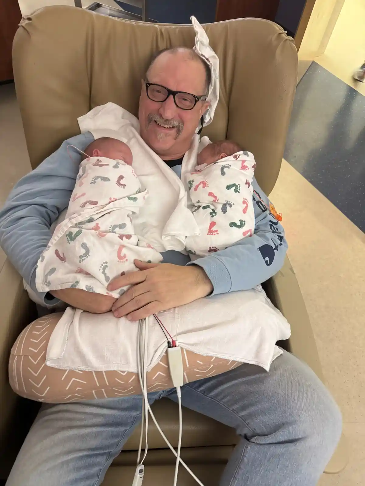 A smiling middle-aged man with glasses and a mustache sits in a beige hospital chair, holding two swaddled newborn babies…