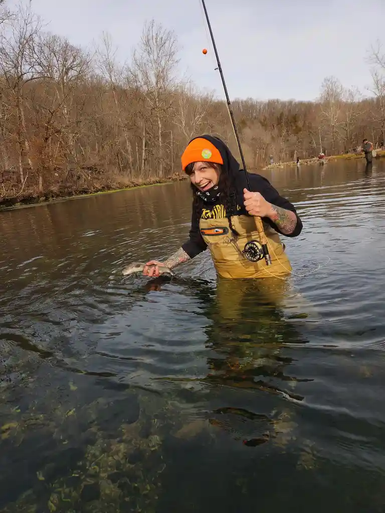 A person wearing waders, an orange beanie, and a black hoodie stands knee-deep in a river holding a small fish in one hand…