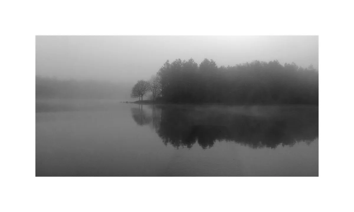 A black-and-white photograph of a misty lake with trees on the far shore, their silhouettes and reflections obscured by fog…