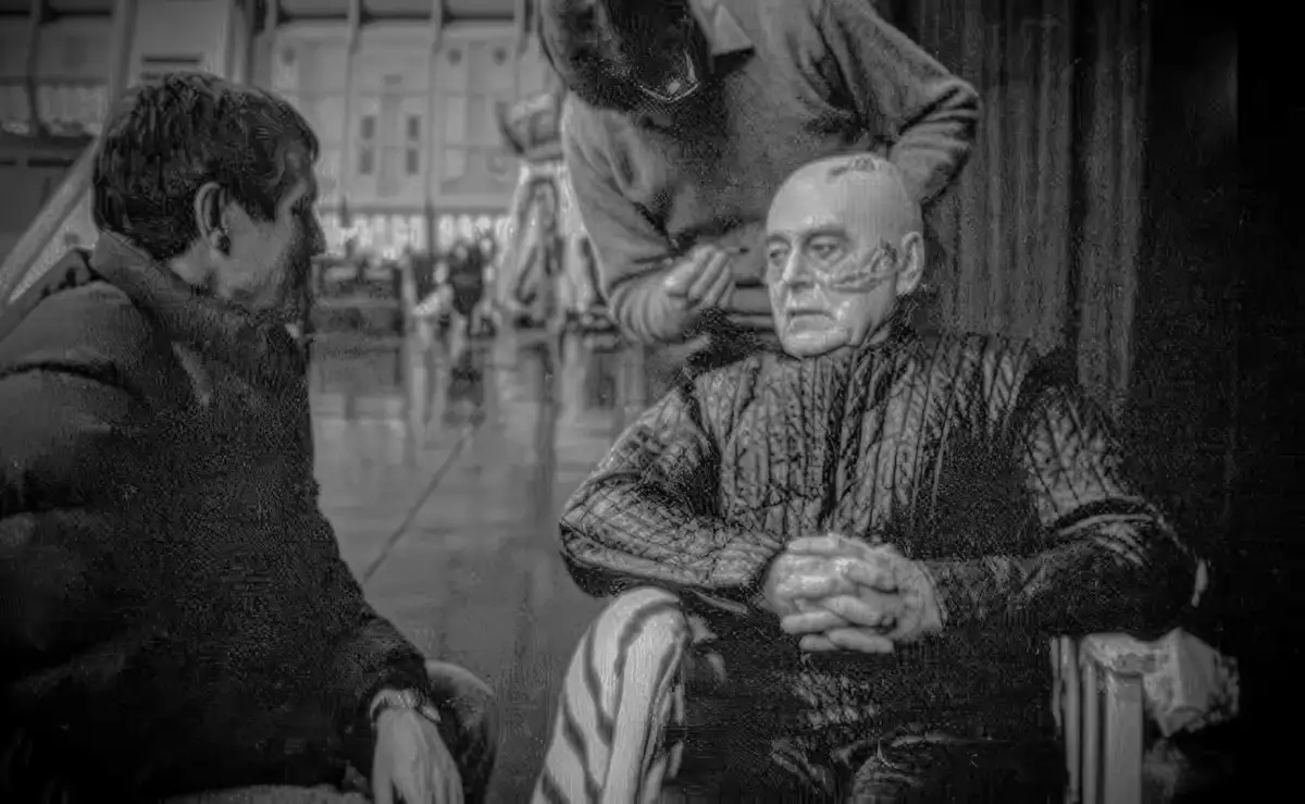 A black-and-white, grainy image shows an older man sitting in a chair while another person applies makeup to his face. A…