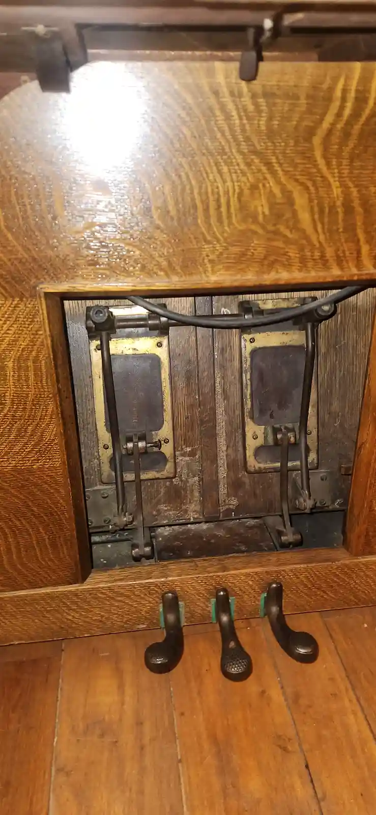 A close-up shot of an old wooden piano's pedal mechanism, showing three metal pedals on the floor and two internal levers…
