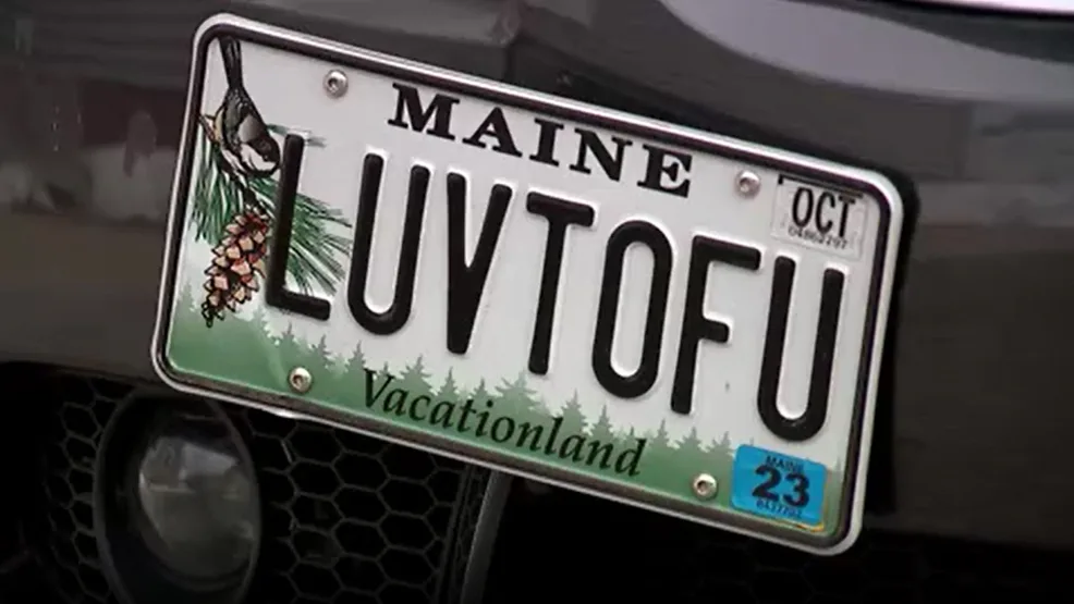 A close-up of a Maine state license plate mounted on a dark vehicle, featuring the text 'LUVTOFU' in large black letters…