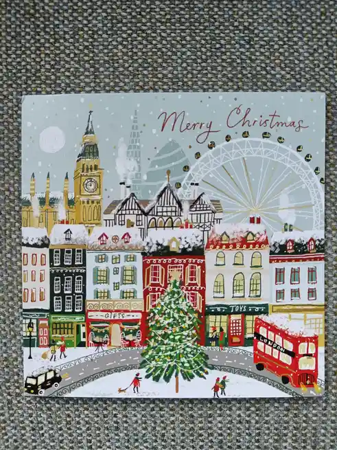 A festive illustrated Christmas card depicts a snowy London street scene with iconic landmarks including Big Ben, the London…