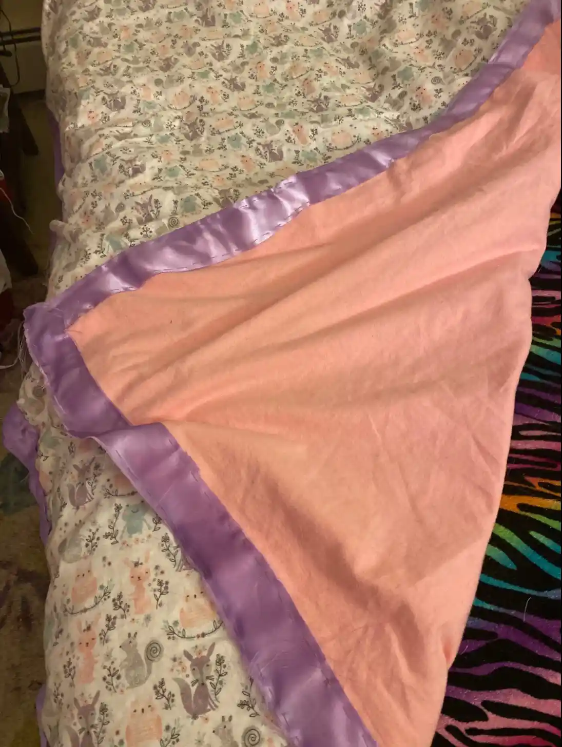 A close-up photo shows a handmade blanket with a soft peach-colored fabric and a shiny purple satin border, laid over a…
