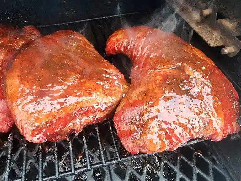 Two large, glistening, sauce-covered meat cuts sizzle on a black grill grate, with smoke rising from the cooking surface…
