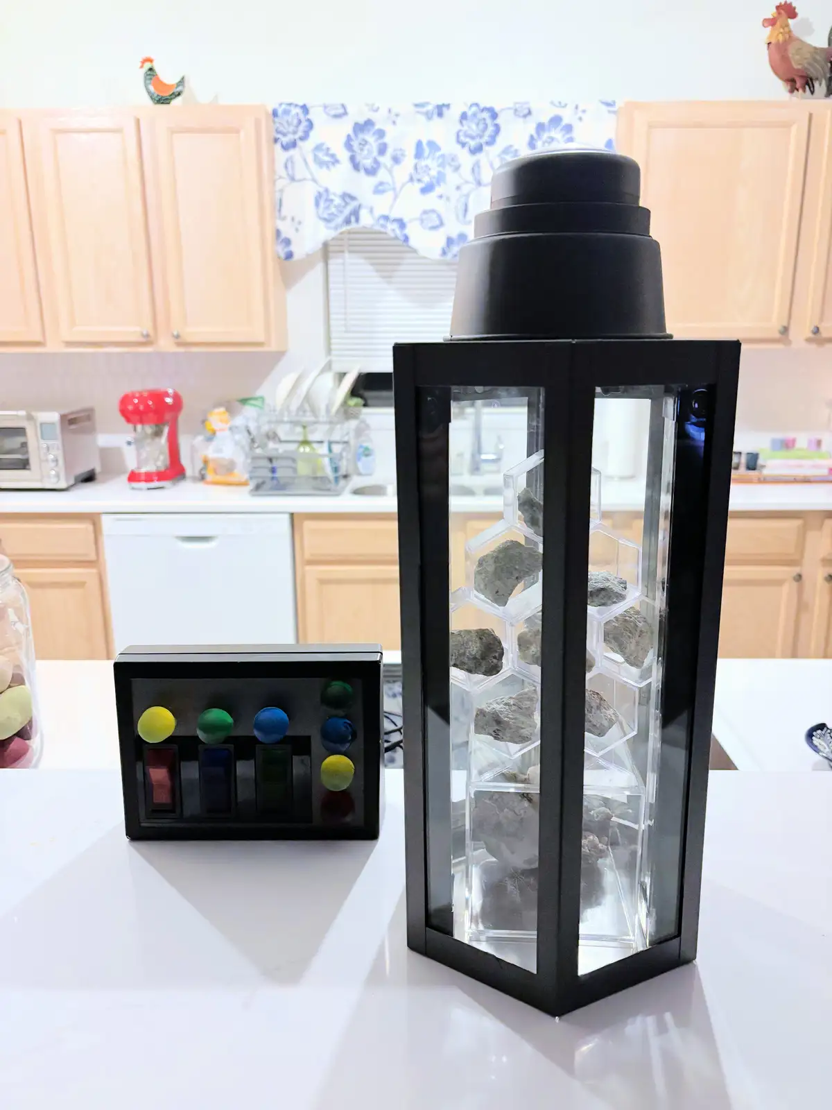A black hexagonal lantern filled with gray rocks sits on a white kitchen counter next to a black remote control with…