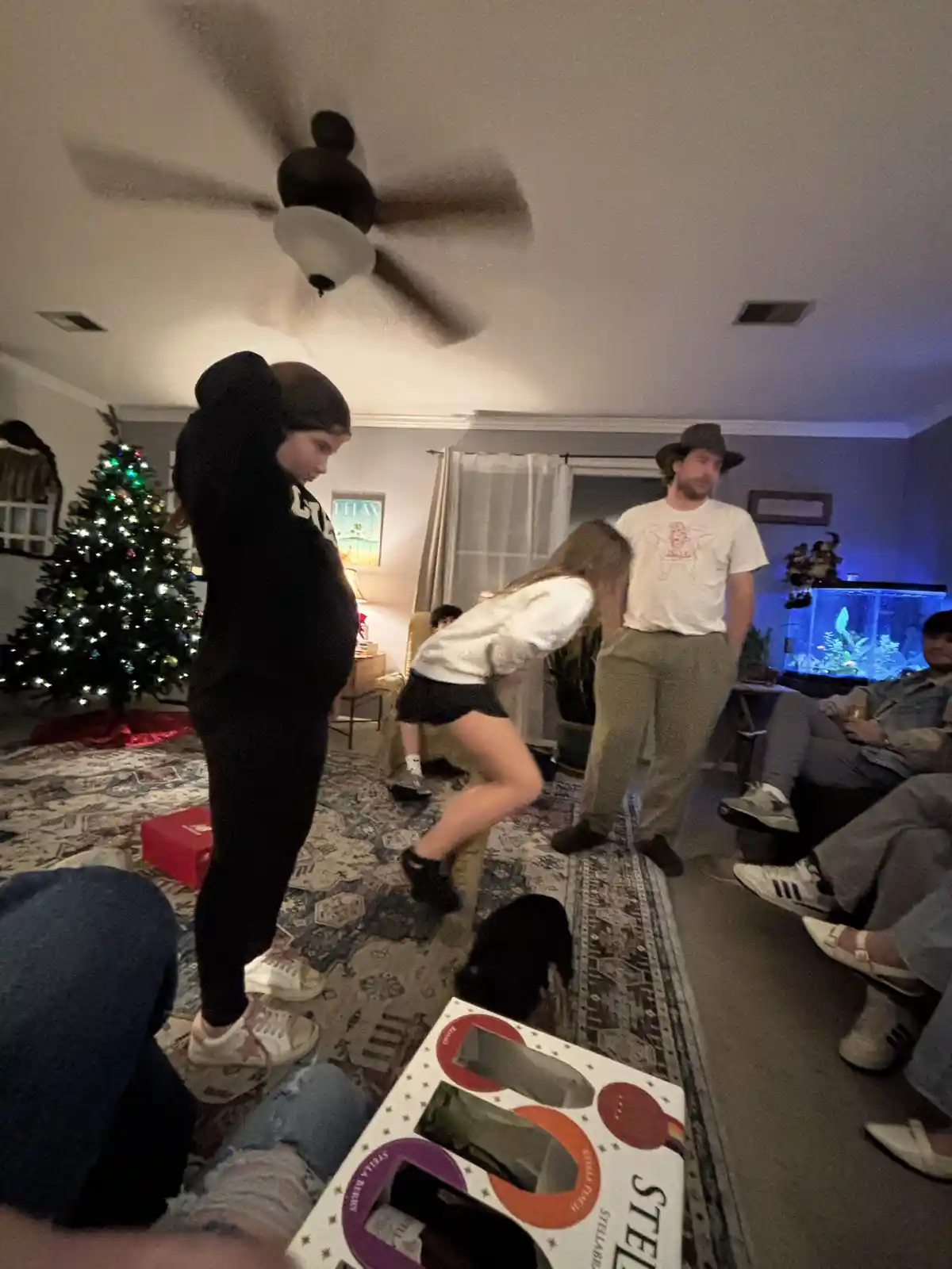 A blurry indoor photo shows a group of people in a living room with a lit Christmas tree in the background. A person in a…