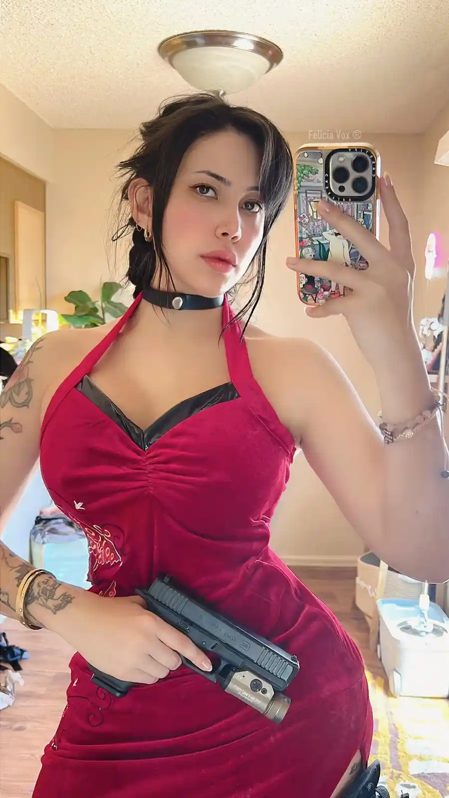 A woman with dark hair and tattoos takes a mirror selfie holding a smartphone with a colorful case, while wearing a red…