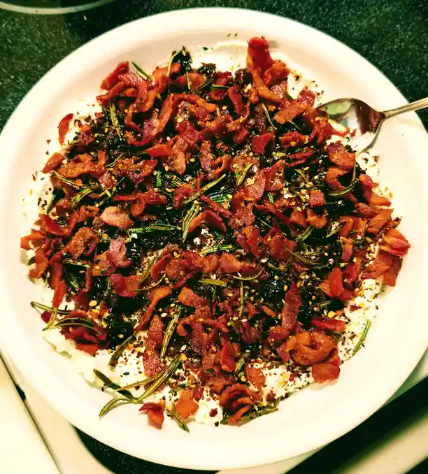 A close-up photo shows a white bowl filled with a creamy white base, generously topped with crispy bacon bits, green…