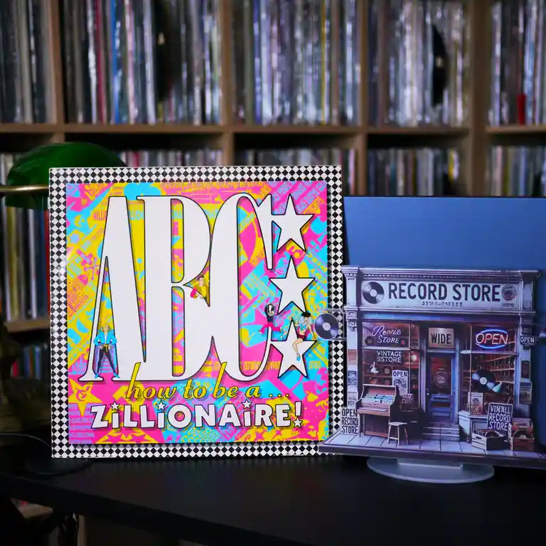 Two vinyl record album covers sit on a desk in front of a wall of records. The left cover is brightly colored with large…